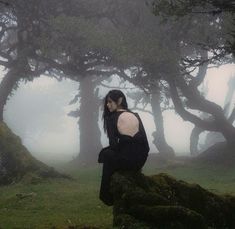 a woman sitting on top of a moss covered rock in front of some tall trees