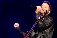 a man holding a guitar in his right hand and singing into a microphone on stage