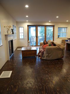 two people sitting on a couch in a living room with wood flooring and large windows