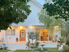 a white house with lots of plants and trees in front of the house at dusk