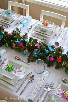 the table is set for christmas dinner with white plates and silver utensils, green garlands, and colorful ornaments