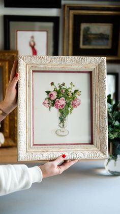 a woman holding up a framed painting with flowers in it's vase on the table