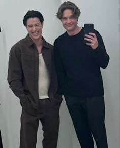 two men standing next to each other in front of a white wall and one holding a cell phone