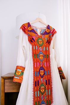 a white dress with colorful designs on it sitting next to a wooden table and window