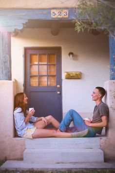 a man and woman sitting on the steps in front of a blue door drinking coffee