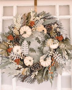 a wreath with white pumpkins, greenery and pine cones hanging in front of a window