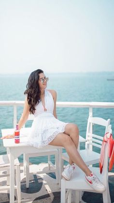 a woman sitting on top of a white bench next to the ocean and holding a drink