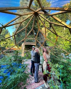 three people standing under a wooden structure surrounded by plants and trees in a garden area