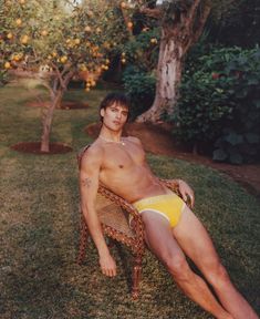 a shirtless man sitting on a chair in an orange tree yard with his legs crossed