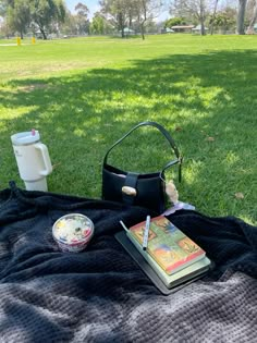 a book, cup and purse sitting on a blanket in the grass near a picnic table