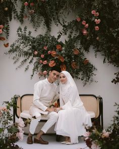 a man and woman sitting on a bench in front of a wall covered with flowers