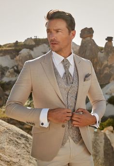 a man wearing a suit and tie standing in front of some rocks with his hands on his hips