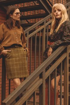 two women standing on the steps of a brick building, one wearing a brown shirt and plaid skirt