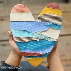 a person holding up a heart shaped piece of paper with mountains and water on it