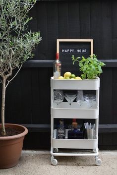 an outdoor bar cart with drinks, lemons and limes on it next to a potted olive tree