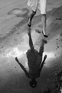 a woman walking across a puddle in the middle of a road with her reflection on the ground