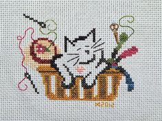 a cross stitched picture of a cat sitting in a basket with scissors and thread