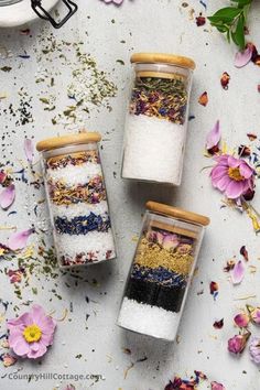 three small jars filled with different types of salts and flowers on a white surface surrounded by petals