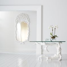 a glass table with flowers on it in front of a white wall and an ornate mirror