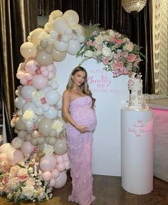 a pregnant woman standing in front of a backdrop with balloons, flowers and a cake