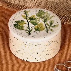 two wedding rings sitting next to a white box with green leaves painted on the lid