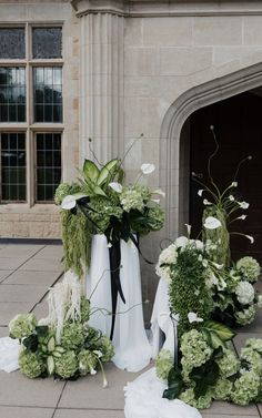 two white vases with flowers and greenery on the ground in front of a building