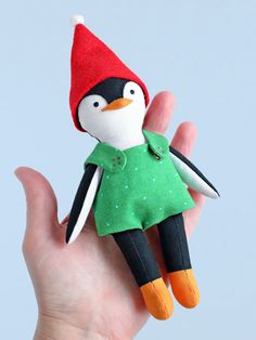 a hand holding a small stuffed penguin wearing a green dress and red hat with an orange beak