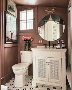 a white toilet sitting next to a bathroom sink under a round mirror on top of a wooden cabinet