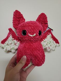 a hand holding a red stuffed animal with wings on it's head and eyes