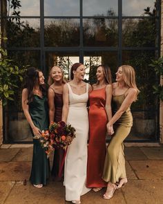 four women standing together in front of a large glass window, all wearing different colored dresses