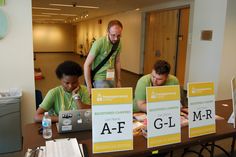 three people are working on their laptops at a table in an office setting with signs that read g i m r