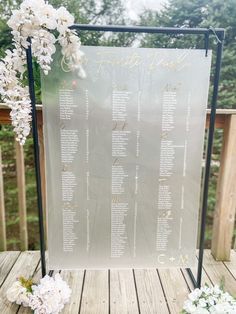 a wedding seating chart with white flowers and greenery on a wooden deck in front of trees