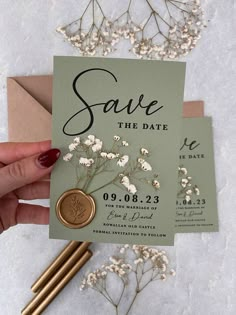 a person holding up a save the date card next to some flowers and envelopes