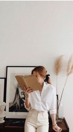 a woman standing in front of a white wall holding a brown book and looking at it