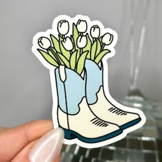 a hand holding up a sticker with flowers in a boot on the bottom and white tulips sticking out of it