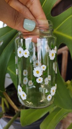 a hand holding a mason jar with daisies painted on the side and green leaves in the background