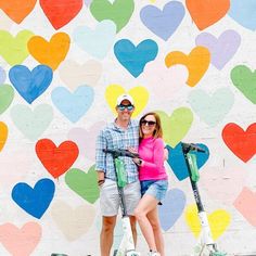 a man and woman standing next to a scooter with hearts painted on the wall behind them