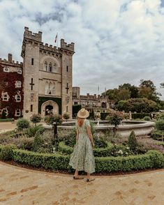 a woman wearing a hat standing in front of a castle like building with a fountain