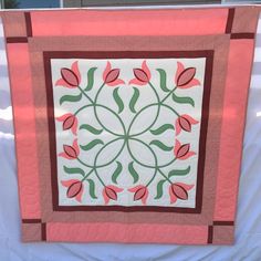 a quilted wall hanging with pink and green flowers on it's side, in front of a window