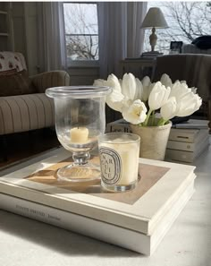 a candle and some flowers on top of a book in front of a couch with a window