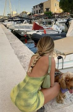 a woman sitting on the dock with her dog next to some boats and buildings in the background