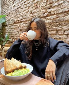 a woman sitting at a table drinking from a cup while holding a sandwich in her hand