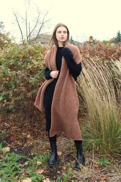 a woman standing in front of some bushes wearing a brown and black coat with long sleeves