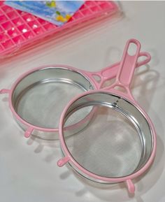 two pink glasses sitting next to each other on top of a white table with plastic containers in the background