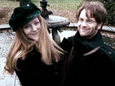 a man and woman standing next to each other in front of a train track with trees