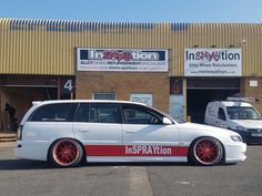 a white car with red rims parked in front of an industrial building next to another vehicle