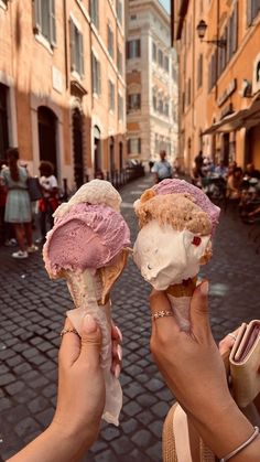 two people holding ice cream cones in their hands on a cobblestone street lined with buildings