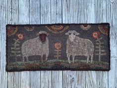 a door mat with two sheep on it and an orange flower in the center, against a wooden background