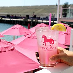 a person holding up a pink pony drink in front of some umbrellas and water