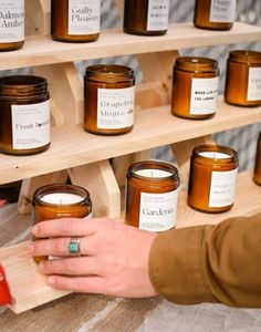 a person holding a candle in front of some jars on a shelf with other candles